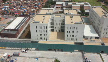 An aerial view of the Hospital Bosa in Colombia. The buildings are white with flat roofs and numerous windows. The complex is surrounded by a green fence, and there are vehicles and additional structures nearby. In the background, a densely populated residential area with colorful houses is visible.