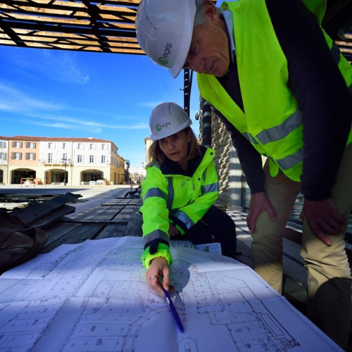 Two construction workers in high-visibility jackets and helmets reviewing architectural plans at a construction site. One worker is pointing at the plans with a pen, while the other looks on. Buildings and a clear blue sky are visible in the background.