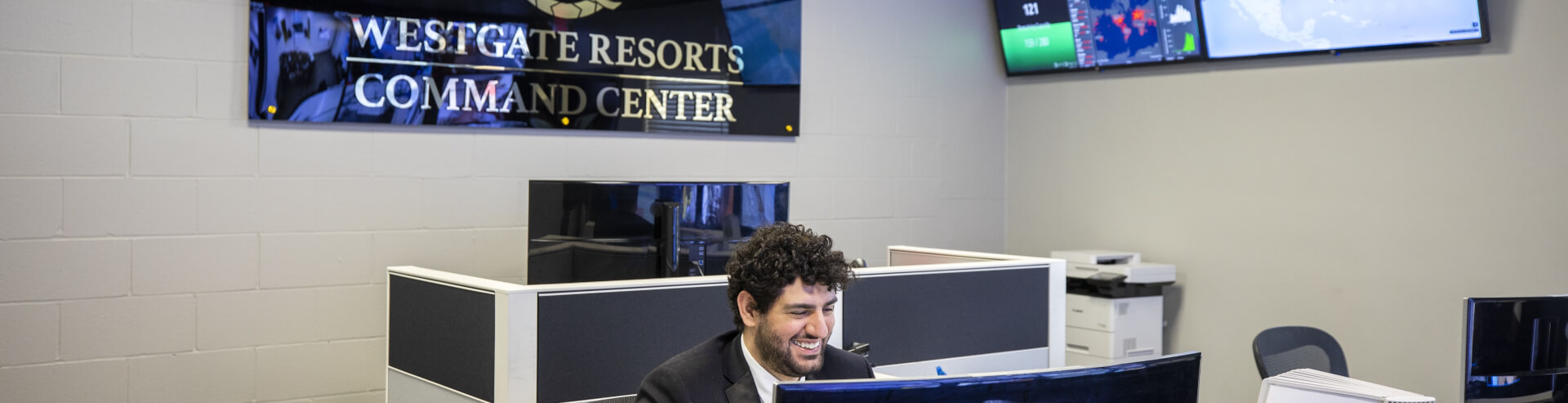 A smiling man sat at a desk on a computer with a sign behind him that reads "westgate resorts command center"