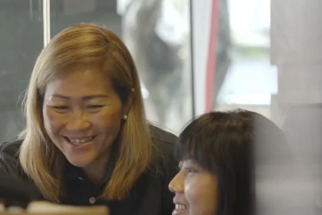 two staff members smiling as one shows the other how to do something on a cumputer