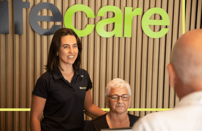Lifecare team member standing beside a seated client in a clinic reception area, with the Lifecare logo displayed in the background. The image represents personalised care, rehabilitation support and collaborative allied health services.