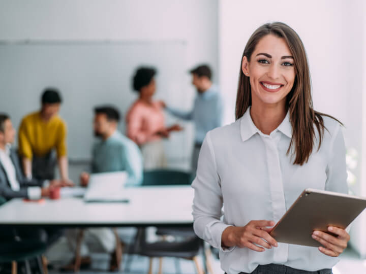 Person holding a tablet in an office environment, with colleagues collaborating in the background, representing professional growth, teamwork and career opportunities within APM's allied health network.