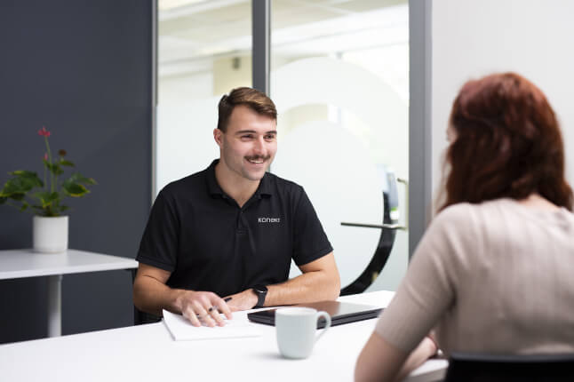 Person seated at a meeting table speaking with another person in a modern office setting. A notebook, tablet and coffee mug are visible, representing collaboration, professional support and client-focused allied health services.