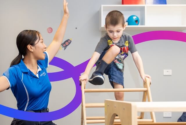 child climbing over climbing frame with therapist watching with hand in the air showing praise and encouragement