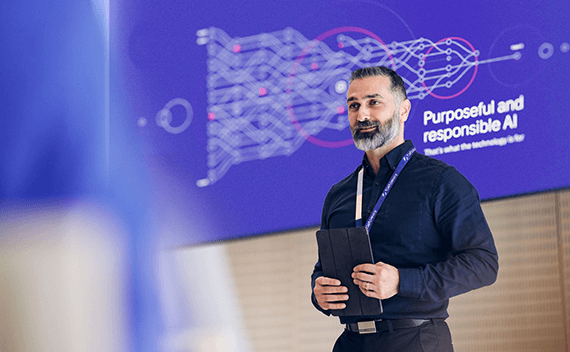 A man giving a presentation, holding a tablet and in front of a big screen