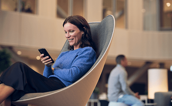 En kvinna med mörkt hår som sitter i en fåtölj, kollar på sin telefon och skrattar