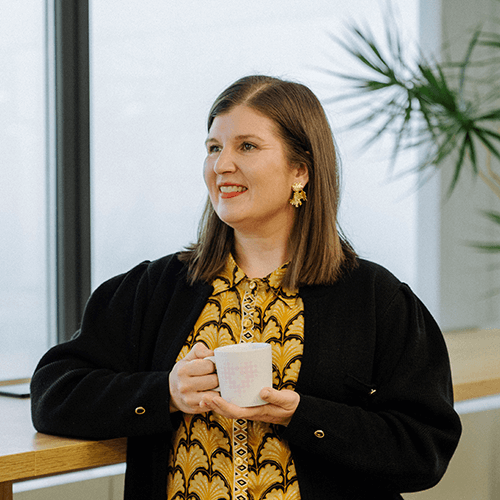 A woman with brown hair, holding her coffee cup and smiling