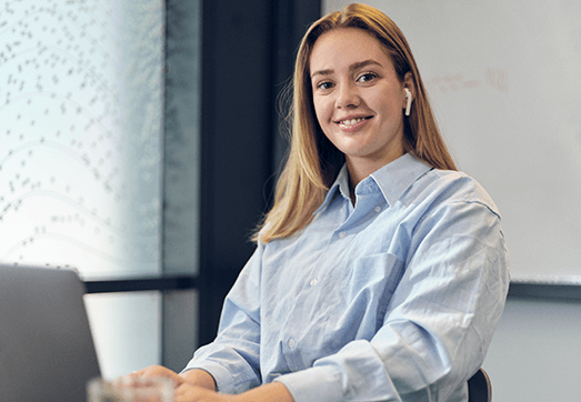 A girl with blonde hair, sitting by her computer with her earphones, smiling to the camera
