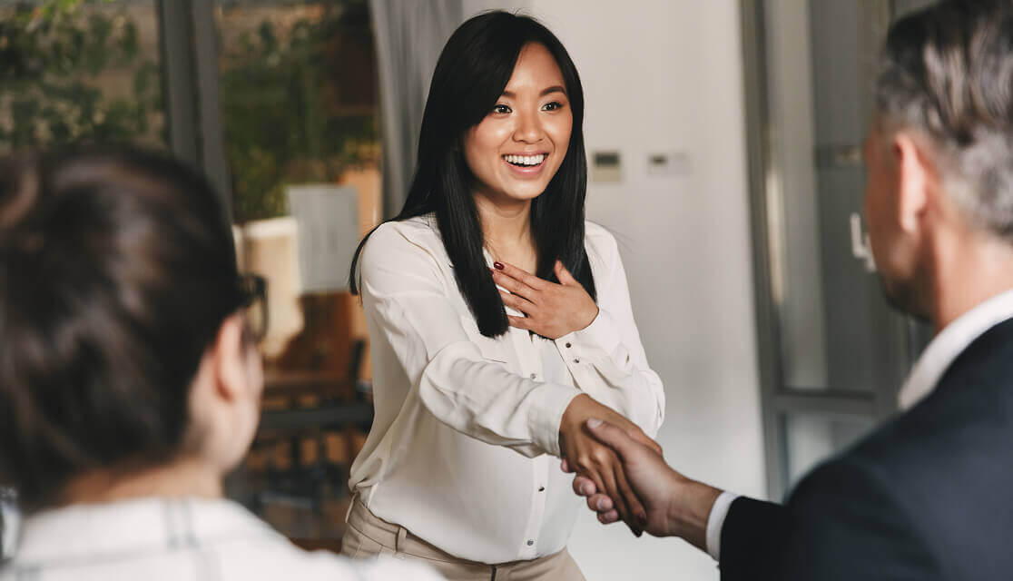 A smiling young woman with her hand on her chest shakes hands with a man in an office setting.