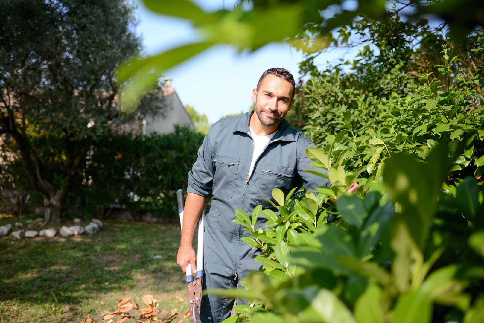 A man in green coveralls smiles while holding a pair of hedge shears and standing next to a leafy bush in a garden.