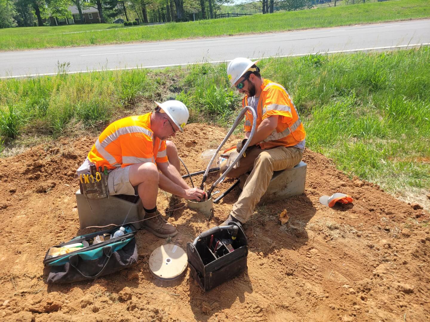 Two utility technicians in orange high-visibility shirts and white hard hats work together on a roadside. 