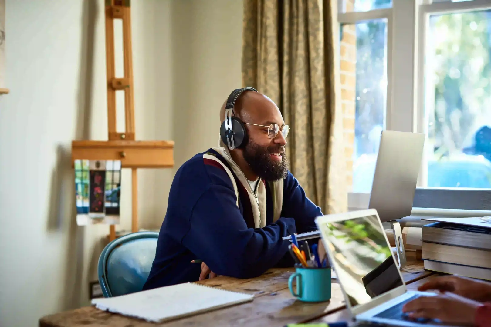 A bearded man wearing headphones and glasses smiles while looking at a laptop screen in a bright home office setting.
